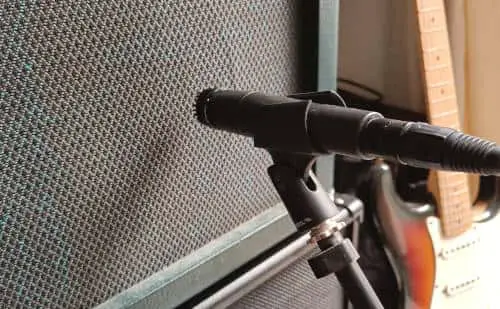 Photo of a microphone in front of a guitar amp for recording, with an electric guitar in the background